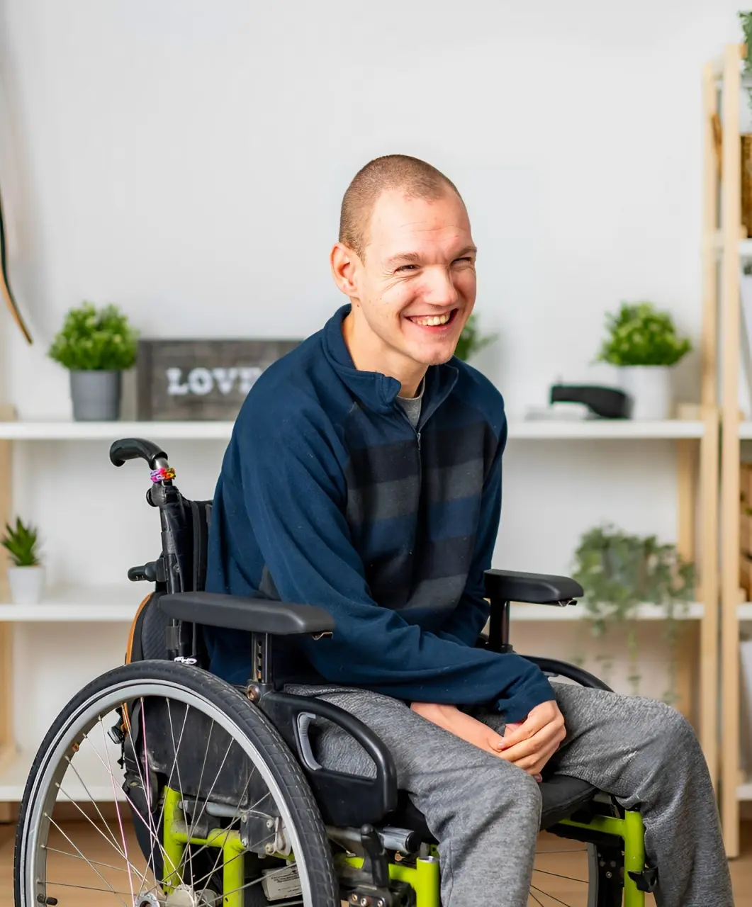 A smiling person in a wheelchair wearing a striped sweater and gray pants is in a room with shelves holding small plants in the background.