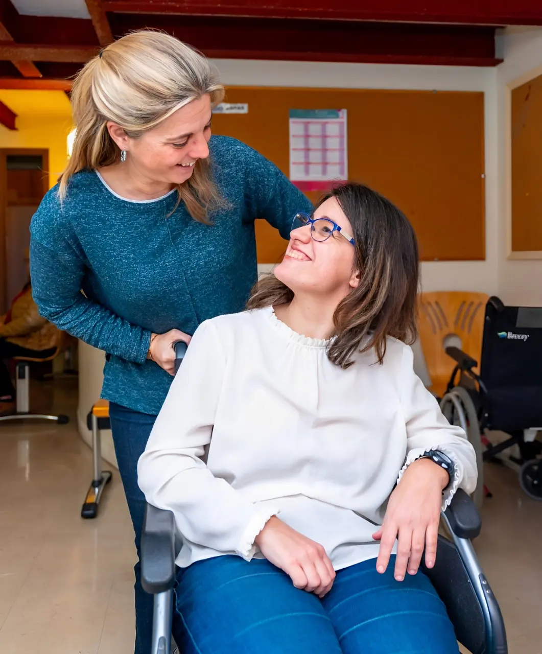 A woman in a blue sweater, representing support services, stands behind and smiles warmly at a woman in a wheelchair indoors. The woman in the wheelchair looks up with gratitude and smiles back.