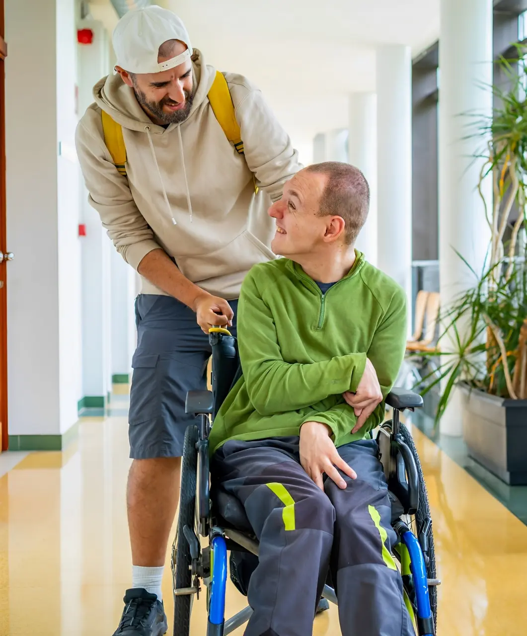 A man pushes another man in a wheelchair down a bright hallway. The man in the wheelchair is smiling at the other man.