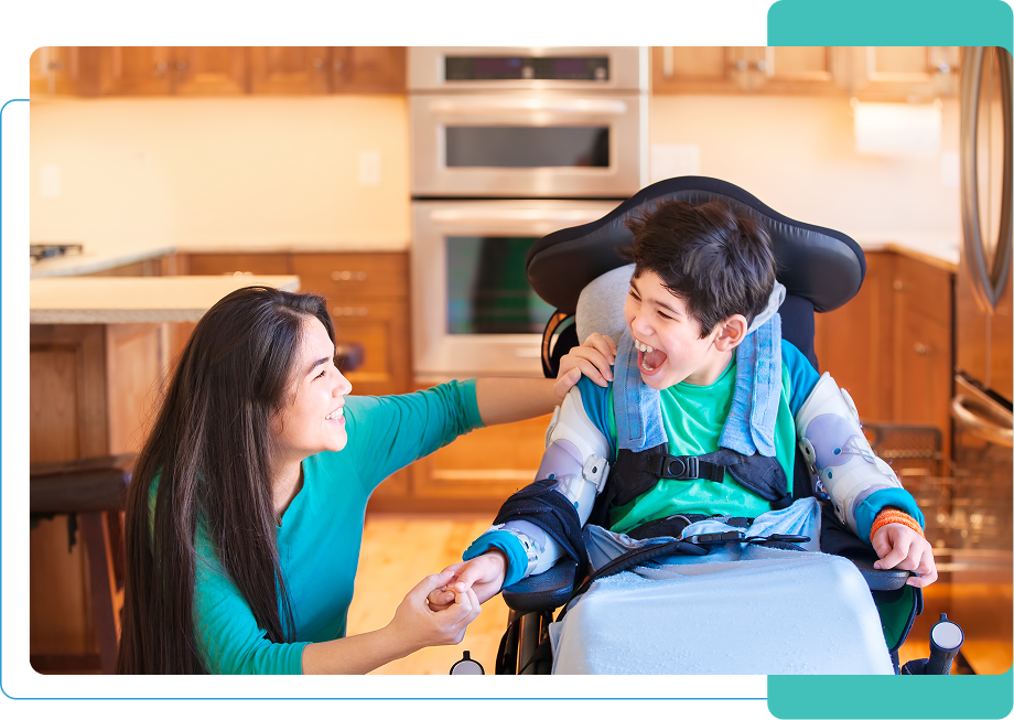 A woman smiles at a laughing child in a wheelchair, in a kitchen filled with love and warmth. The child, wearing supportive gear, clutches her hand as they share a moment under the gentle glow of support services enriching their day.