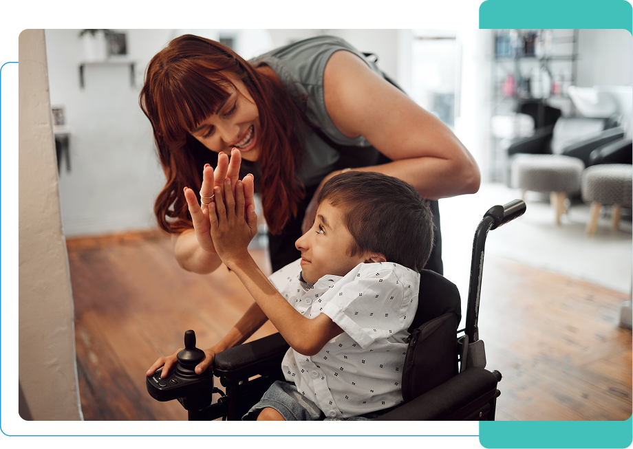 A woman leans down to give a high-five to a smiling child in a wheelchair inside a room with wooden floors.