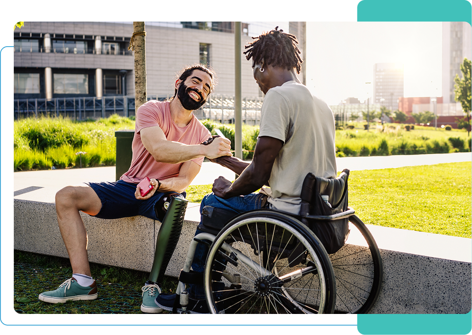 Two men outdoors shake hands warmly. One has a prosthetic leg and holds a beverage, while the other is in a wheelchair. They sit on a concrete ledge with a green park and buildings in the background.