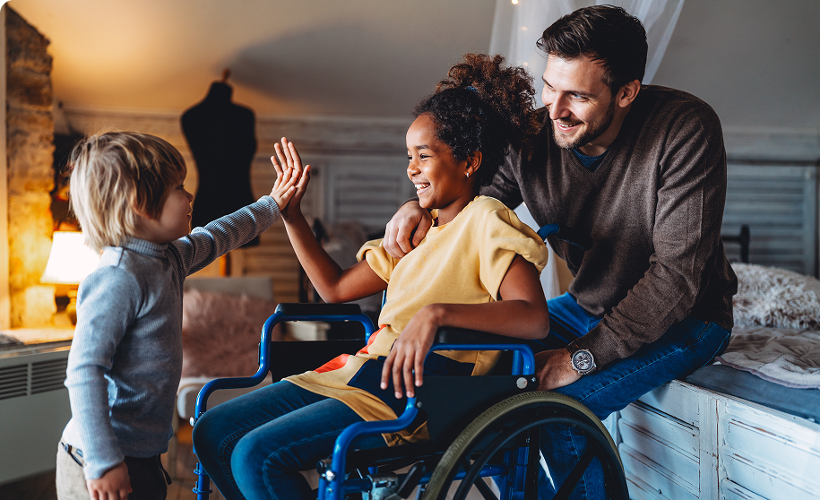 A man smiles at a girl in a wheelchair, sharing a high-five with a standing young boy. The softly lit room exudes a cozy ambiance, reflecting the spirit of support coordination services that bring warmth and connection.