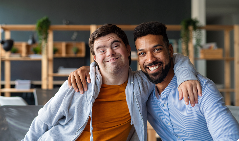 Two men smiling and sitting together in a modern office space, with one arm around each other, embody the spirit of support coordination services.